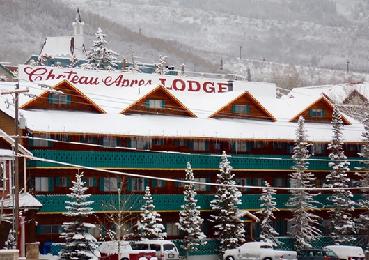 Snow-covered Chateau Apres Lodge with pine trees in front offers cozy private room rentals, nestled in a mountainous area. Cars are parked outside, and everything is blanketed in snow.