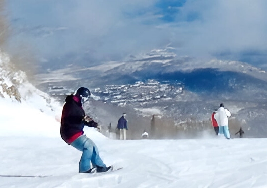 A person gracefully snowboards down the slope, while skiers glide in the background. The stunning snowy landscape and majestic mountains unfold beneath a cloudy sky, perfect for those seeking thrilling adventures followed by cozy evenings in nearby private room rentals.