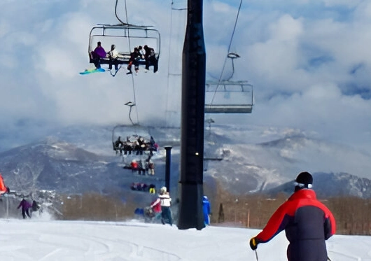 Skiers and snowboarders gracefully carve down the snowy slope as several people enjoy the chairlift in the background. Majestic mountains and clouds provide a stunning backdrop, perfect for those seeking nearby private room rentals to complete their alpine adventure.