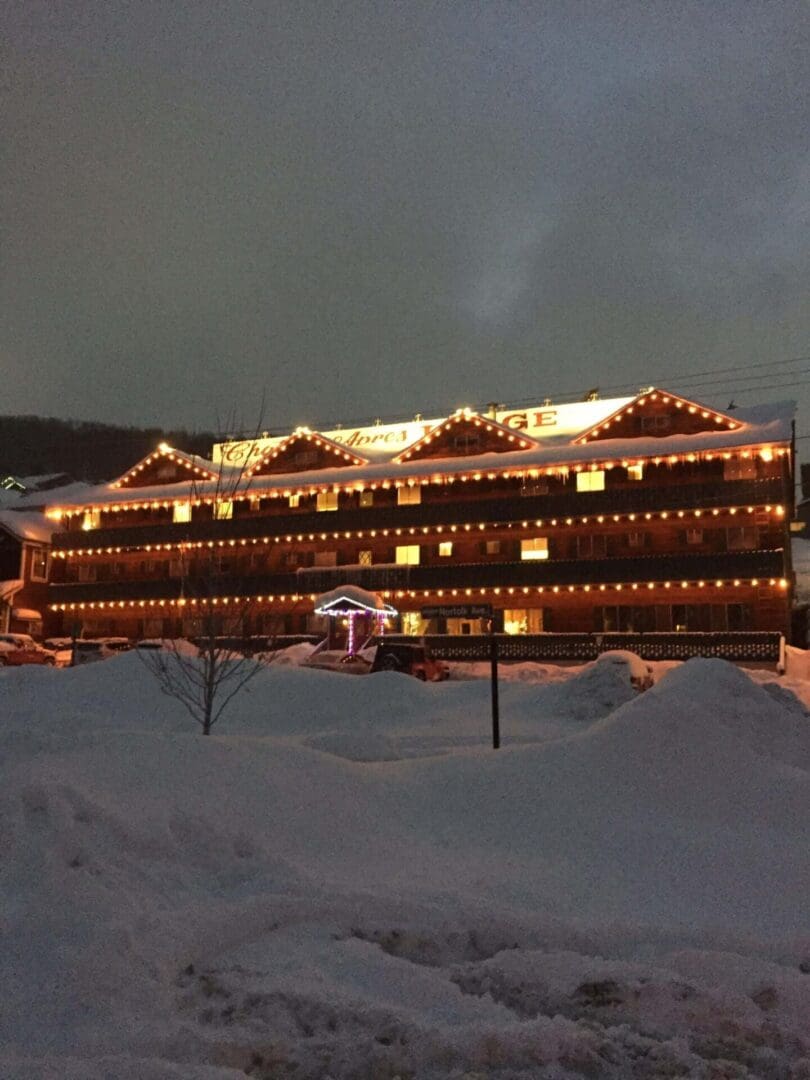 A building with multiple balconies is decorated with white and yellow string lights. Snow covers the ground, and a nearby structure is also lit with purple lights.