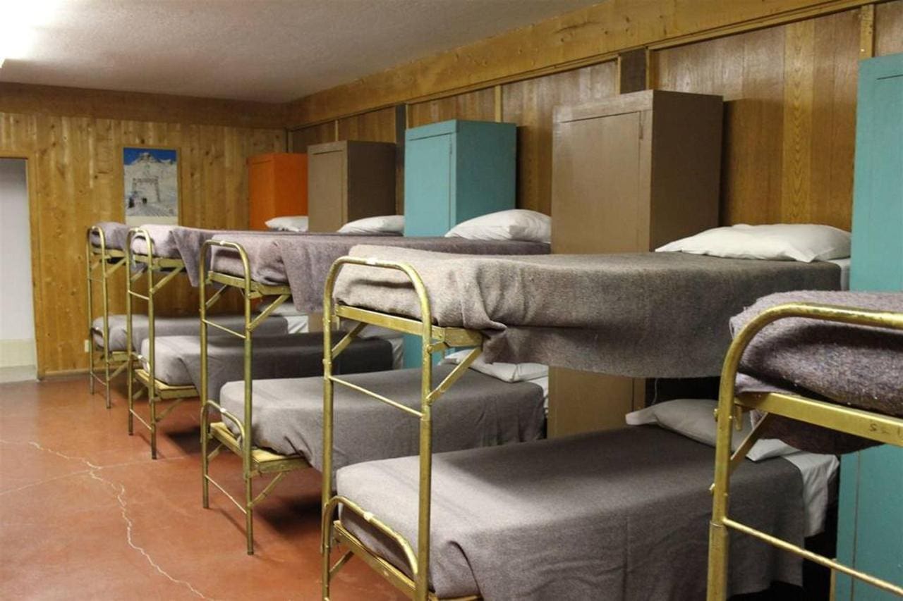 A lodge featuring dorm rooms with a row of metal bunk beds, each outfitted with gray blankets and pillows. The room boasts wooden paneled walls and lockers at the end of each bed.