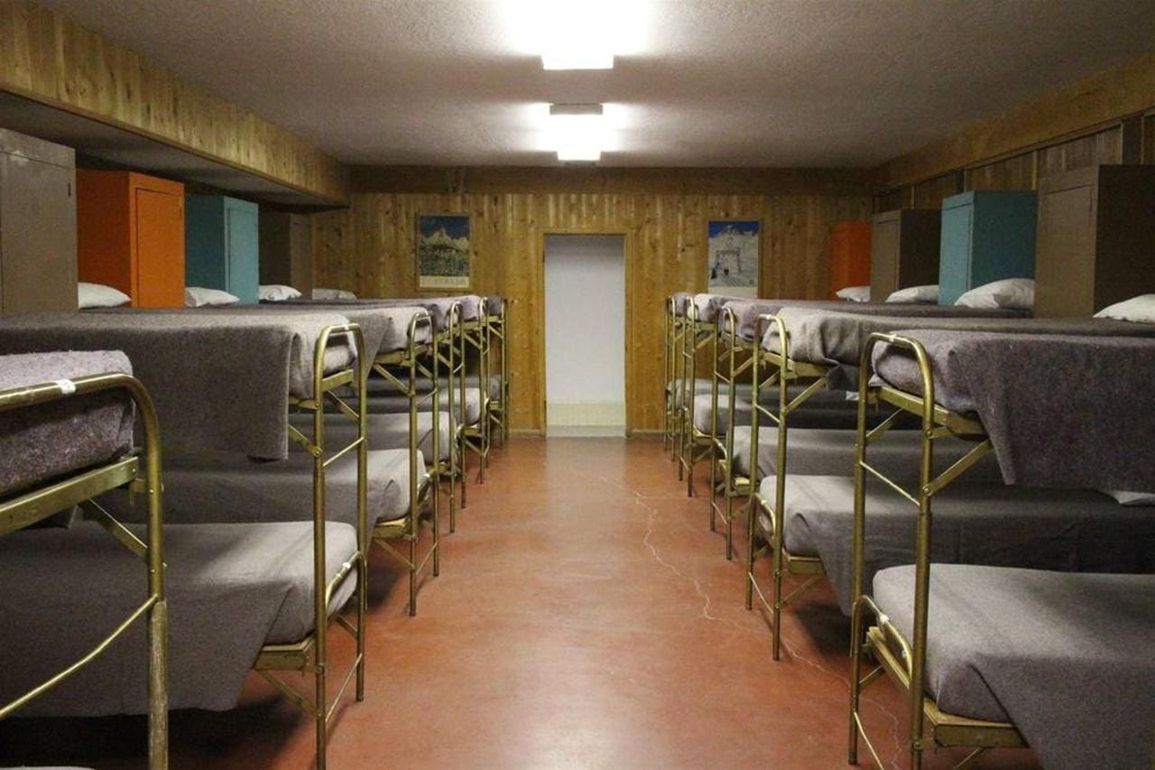 A lodge with dorm rooms featuring multiple bunk beds, each draped in gray blankets and neatly arranged in rows on a polished floor beneath fluorescent ceiling lights.