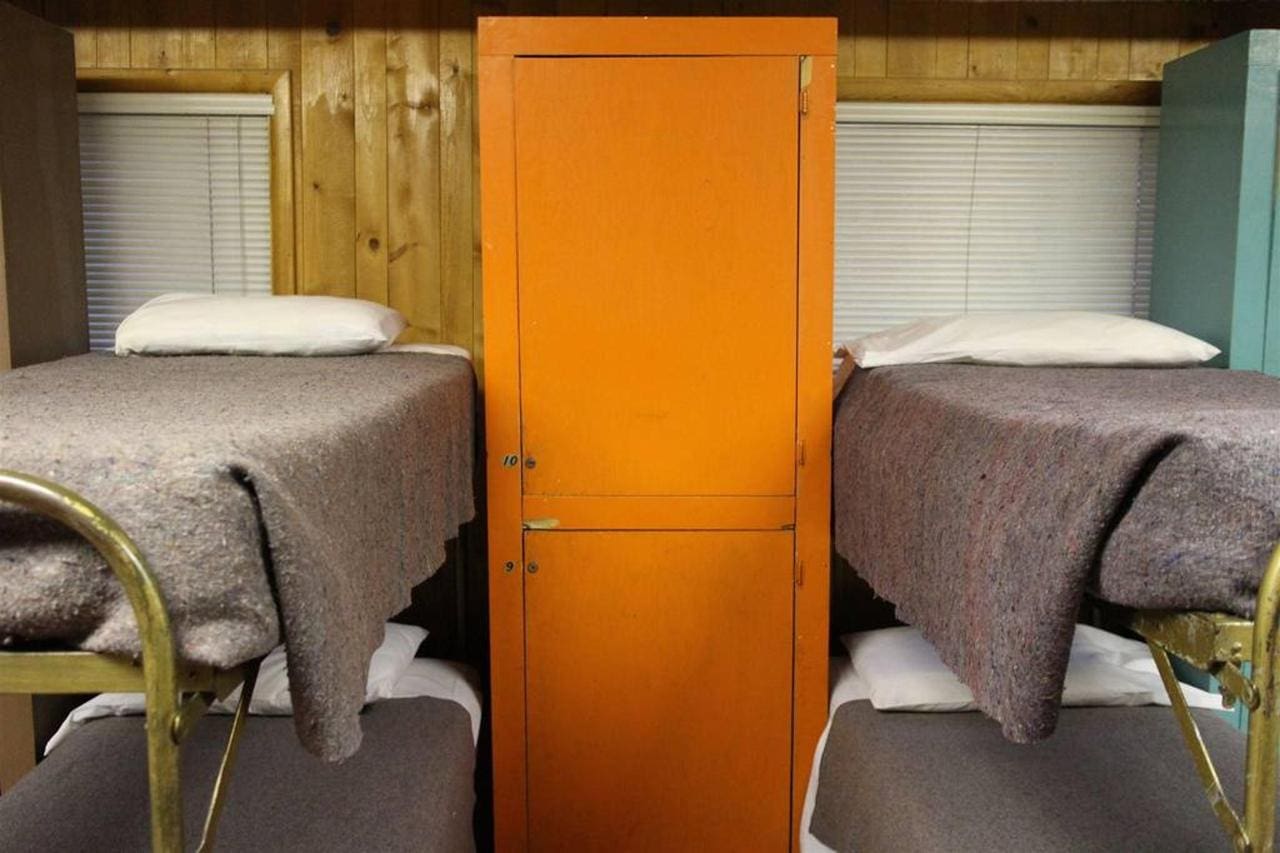 A lodge with dorm rooms featuring two bunk beds on each side and an orange locker at the center, surrounded by wood-paneled walls and blinds on the windows.