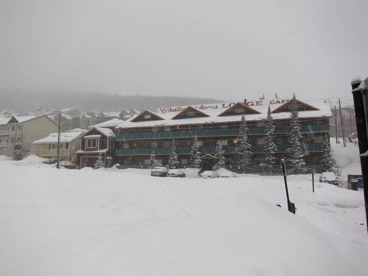 Snow-covered lodge surrounded by snowdrifts with evergreen trees in front. Overcast sky in the background.