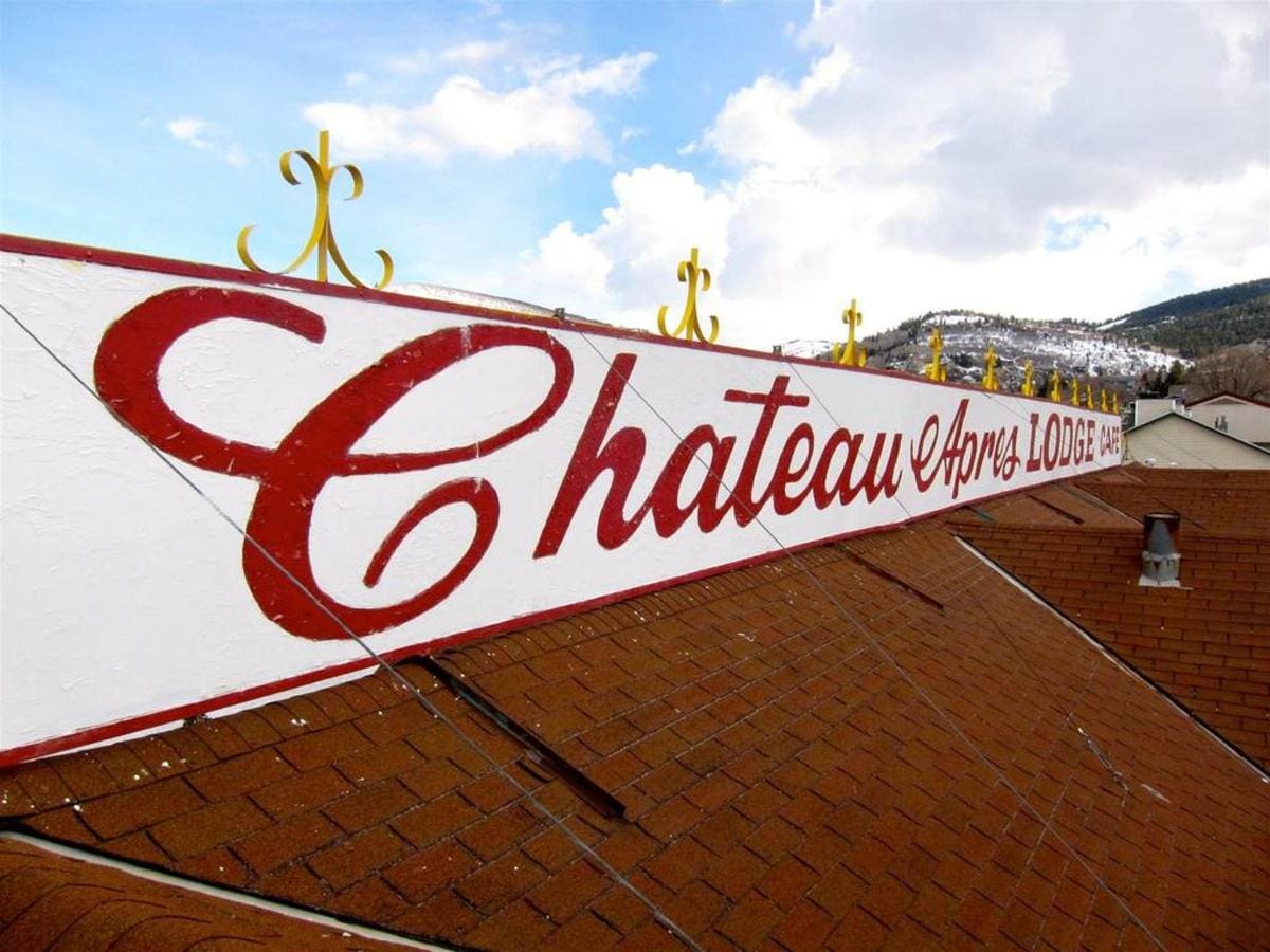 Red and white sign on a roof reads "Chateau Apres Lodge" with decorative trim and a mountain backdrop.