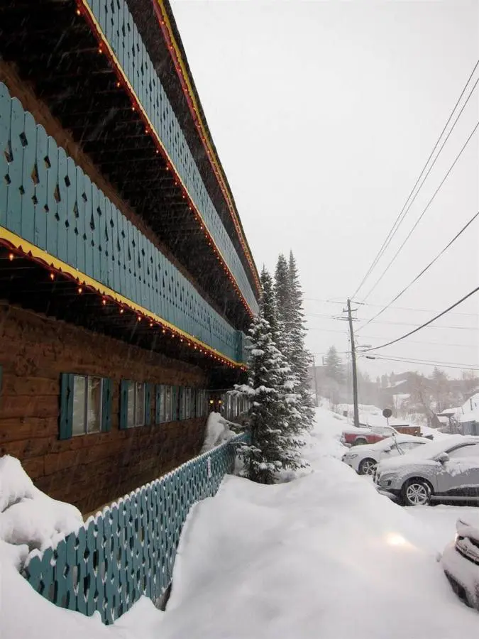 Snow-covered street next to a building with teal wooden balconies and lights. Pine trees and parked cars are visible under overcast skies.