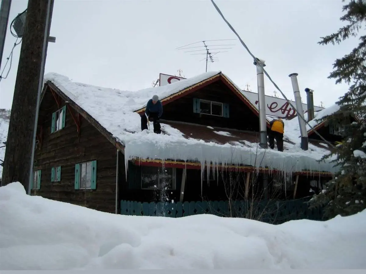 Two people are clearing snow from the roof of a wooden house with icicles hanging from the edge.