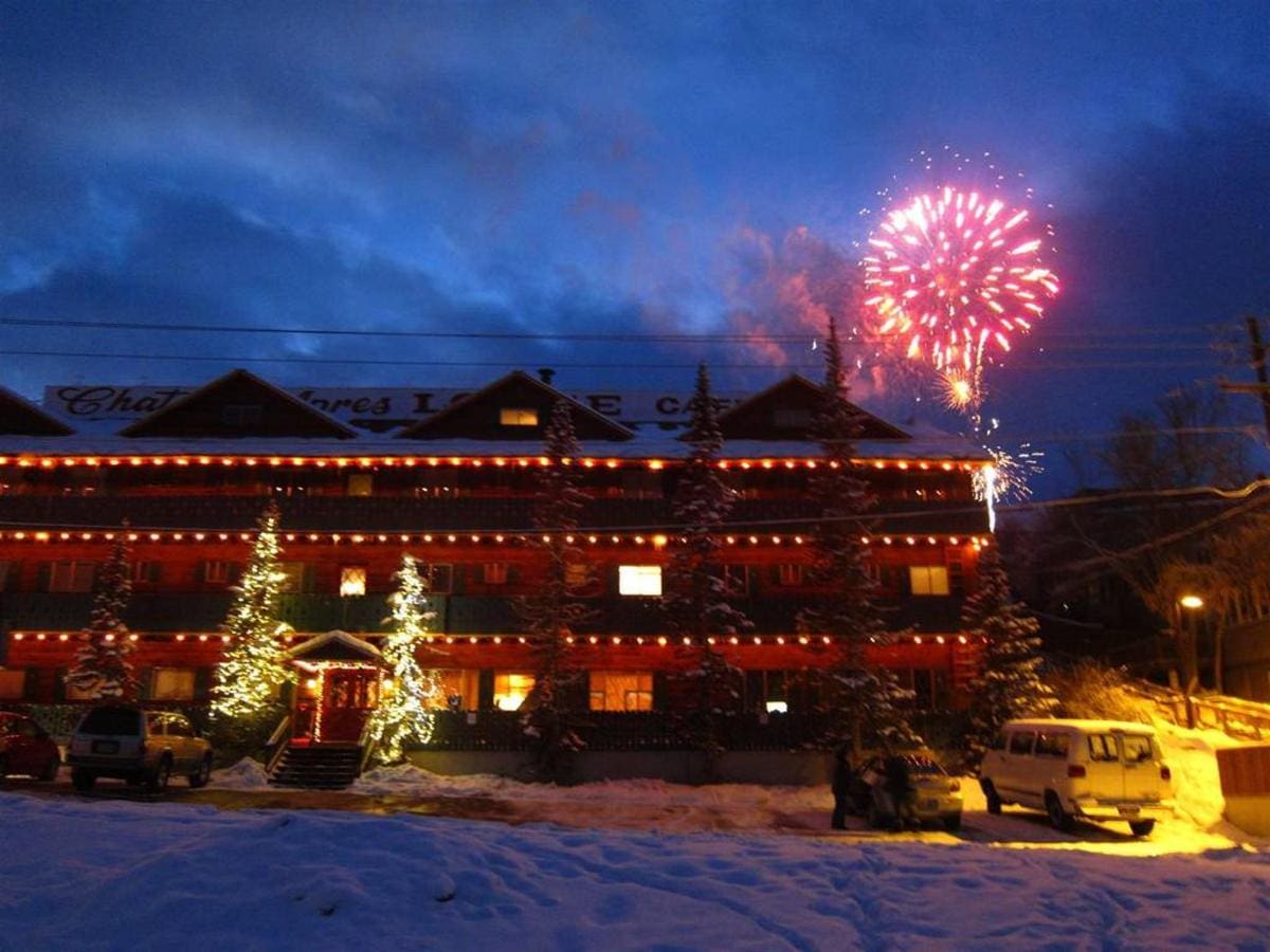 Fireworks light up the sky above a lodge decorated with holiday lights, surrounded by snow-covered trees and a parked van at dusk.