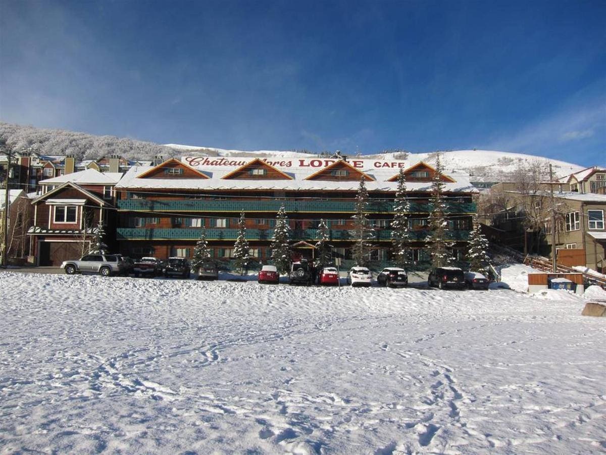 Snow-covered lodge with multiple cars parked in front, surrounded by snowy hills under a clear blue sky.