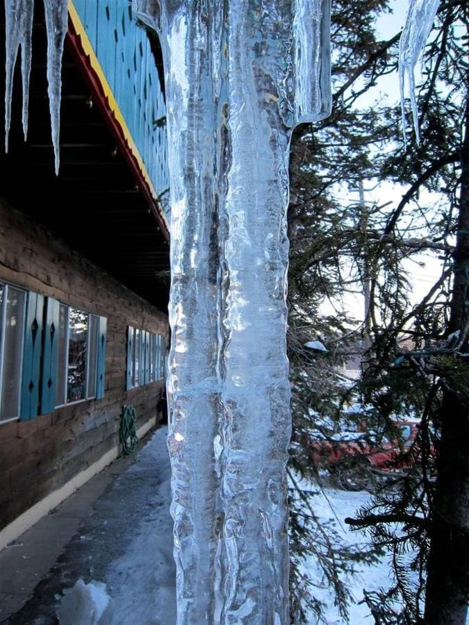 A large icicle hanging from the roof edge of a building, with a wooden wall and snowy ground visible, set against a backdrop of bare trees in winter.