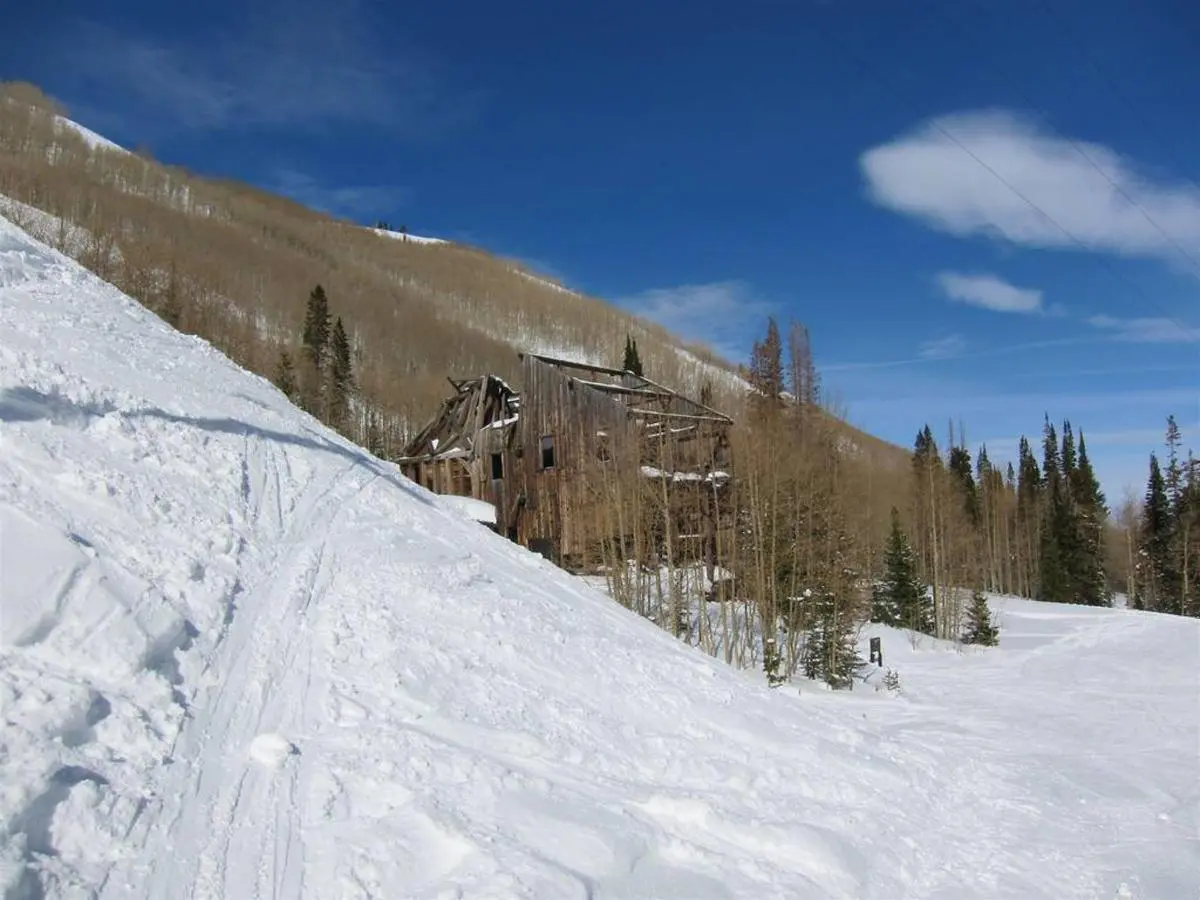 A dilapidated wooden building stands on a snowy slope surrounded by leafless trees under a clear blue sky, embodying the rustic charm of Park City Ski Vacation Rentals.