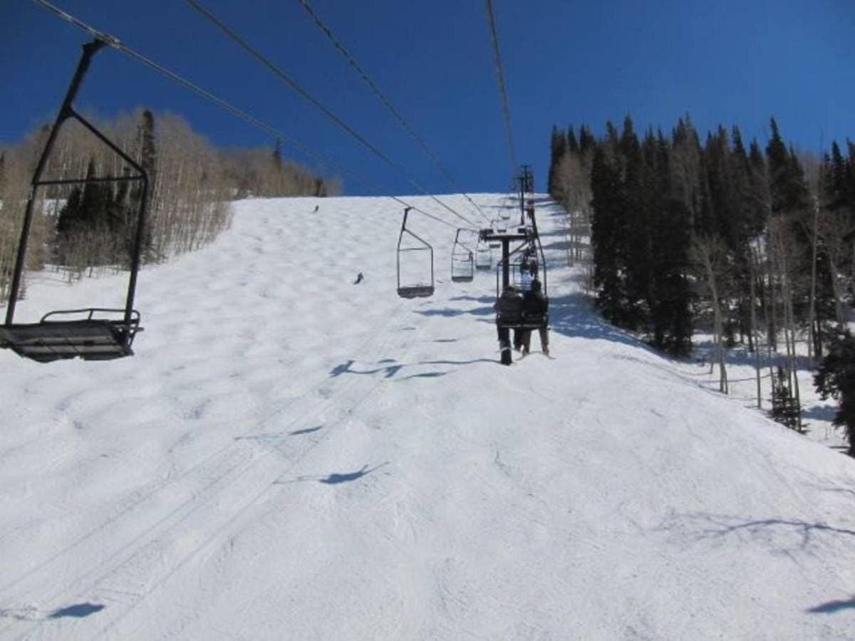 Ski lift ascending a snowy slope with scattered trees and a skier on the hill, all beneath a clear blue sky—a perfect scene for those enjoying Park City ski vacation rentals.