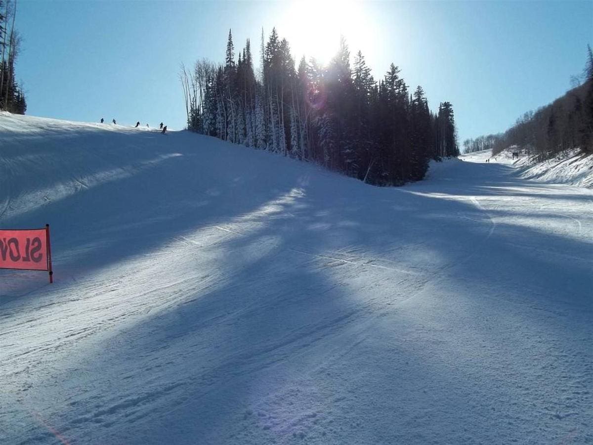 A snowy ski slope in sunlight, surrounded by a forested area with a red warning sign, offers a perfect view for those enjoying their Park City Ski Vacation Rentals.