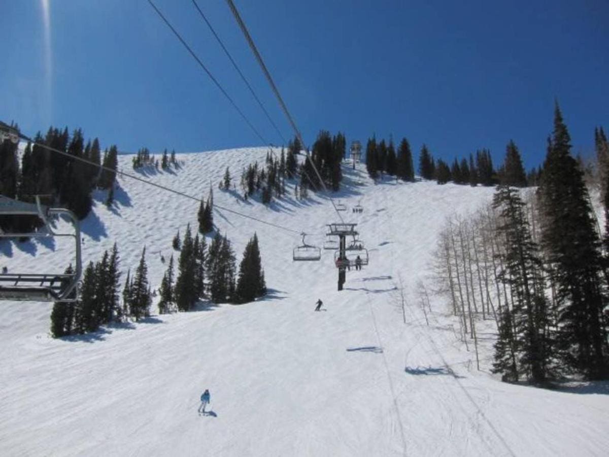 Skiers glide down a snowy slope under a clear blue sky, framed by trees and the gentle ascent of a ski lift. Experience this winter paradise with Park City Ski Vacation Rentals, your gateway to an unforgettable alpine adventure.