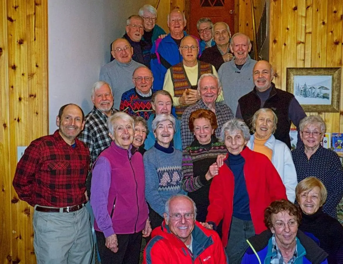 A group of elderly people pose together indoors, with wooden walls in the background, smiling at the camera. They're enjoying their getaway, made even more special by a free bus route that conveniently brought them to this cozy lodging, where good company and laughter fill the air.