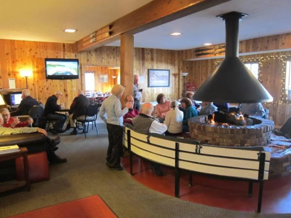 People seated around a central fireplace in a cozy, warmly lit room with wooden walls enjoy the comfort of their lodging. A TV is mounted on the wall, offering entertainment after a day exploring via the free bus route.