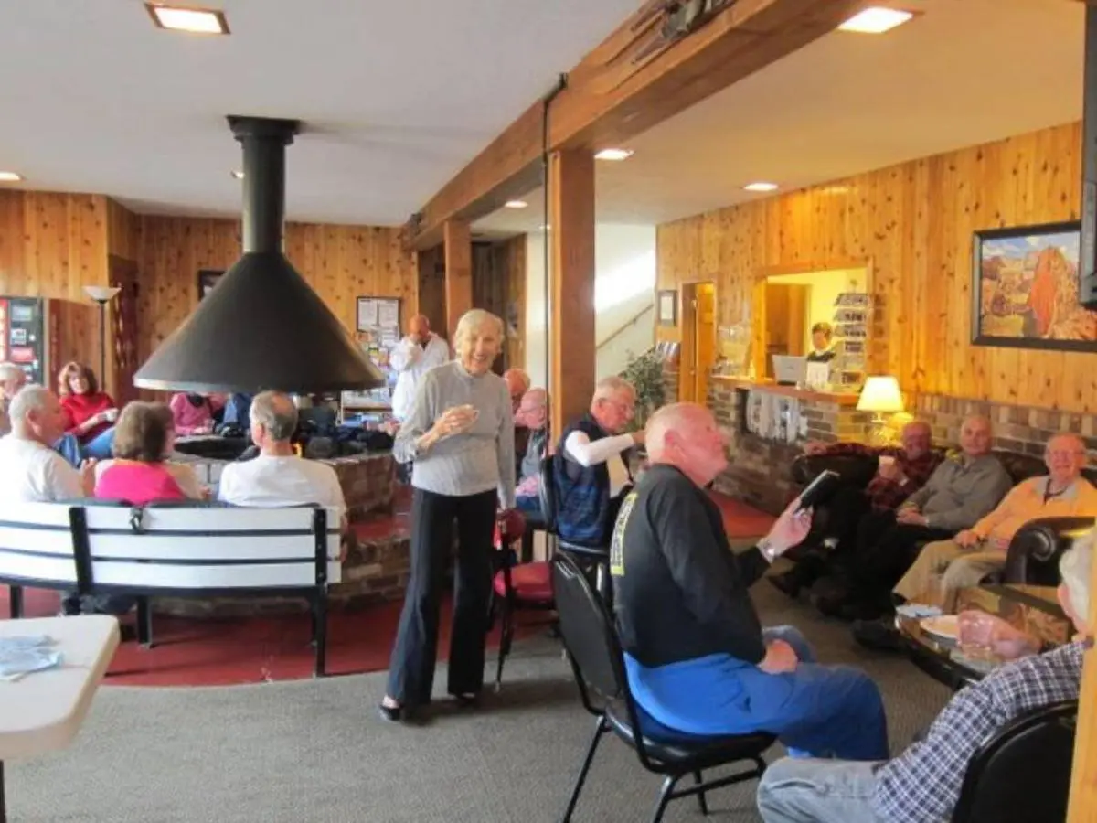 A group of elderly people socializing in a cozy room with wooden walls, seated on chairs and benches. A large fireplace is in the center, providing warmth after returning from exploring courtesy of the free bus route offered by their lodging.