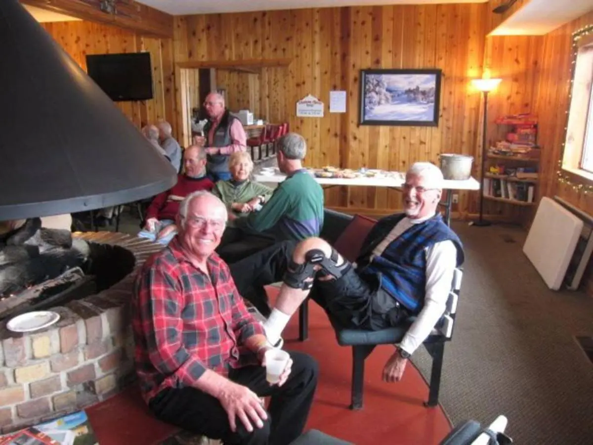 In a cozy wooden room with a fireplace, friends gather around a table, sharing smiles and stories. One man, sporting a leg brace, seems comfortable knowing the nearby free bus route makes their peaceful lodging easily accessible for all.