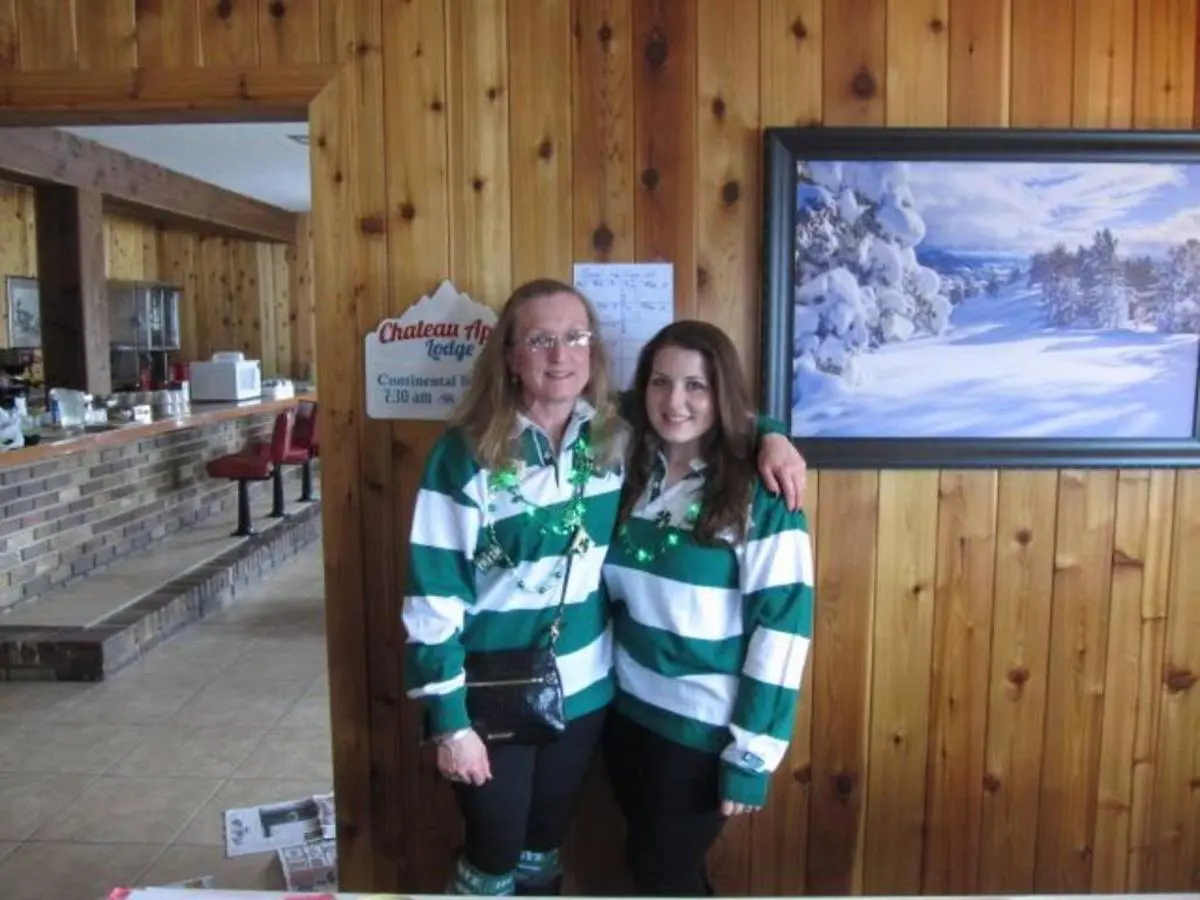 Two people wearing matching green and white striped shirts stand in a wooden room, reminiscent of cozy lodging, beside a snow landscape painting.