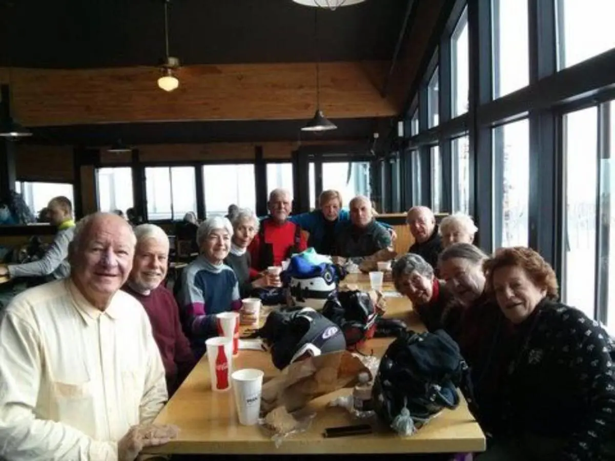 A group of elderly people sitting around a wooden table in a restaurant, smiling towards the camera, enjoying their day out thanks to the free bus route. Drinks and bags are scattered on the table, capturing a moment filled with joy and companionship.