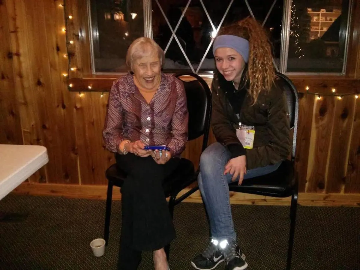 An elderly woman and a young woman sit in chairs, smiling in a cozy wood-paneled room adorned with string lights, sharing stories about their travels on the free bus route.