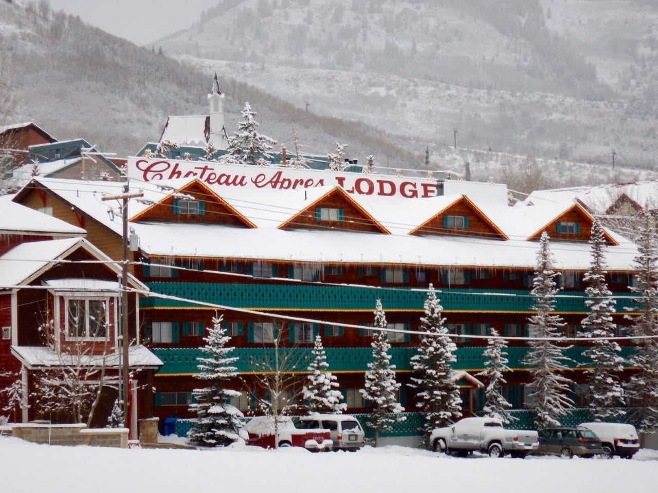 A snowy lodge with green accents and a sign reading "Chateau Aspen Lodge" in a mountainous winter setting.