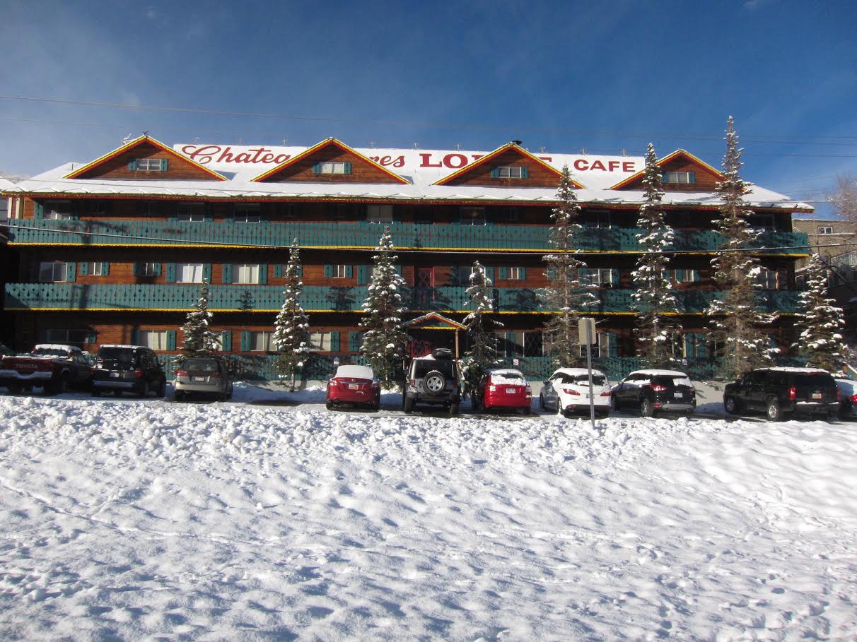 A snow-covered lodge with several parked cars in front, and signs for "Chateau," "Lodge," and "Cafe" on the roof. Pine trees are in the foreground.