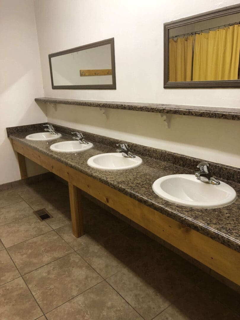 Bathroom sinks with four basins and faucets are set in a long granite countertop, perfect for a lodge with dorm rooms. A mirror stretches above the countertop, reflecting the charm of the yellow curtain in the background. The beige tiled floor adds to the rustic yet cozy atmosphere.