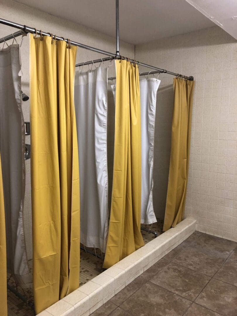 A row of shower stalls with yellow curtains on metal rods lines the tiled bathroom, reminiscent of a lodge with dorm rooms for travelers.