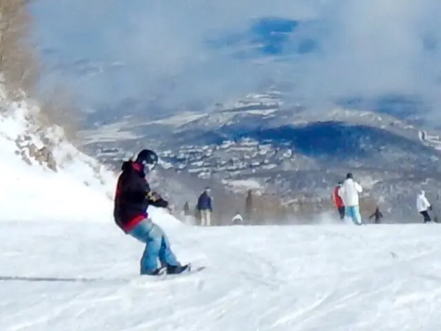 A person snowboards down a snowy slope, surrounded by others enjoying the thrill. The bright, cloud-dappled sky and stunning mountain landscape epitomize a Park City ski vacation.