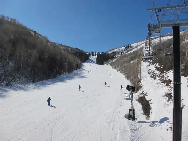 Skiers glide down a wide, snow-covered slope flanked by trees in Park City. A ski lift runs alongside, with a bright blue sky overhead, making it the perfect day to enjoy your ski vacation rental.