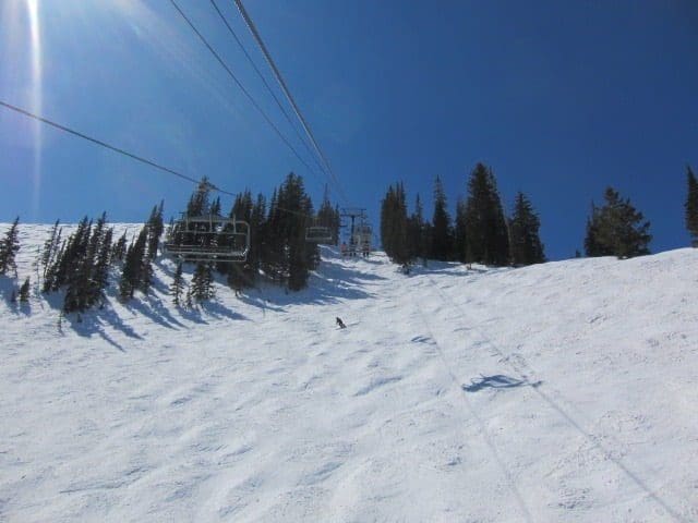 The ski lift ascends a snowy mountain slope with scattered trees under a clear blue sky, while one person glides down, enjoying the pristine conditions. Discover your perfect getaway with Park City Ski Vacation Rentals and experience this winter wonderland firsthand.