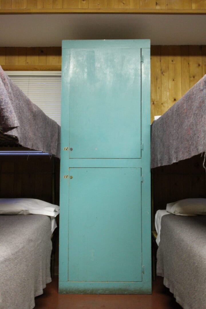 A teal cabinet stands between two bunk beds with gray blankets in a wood-paneled lodge dorm room.