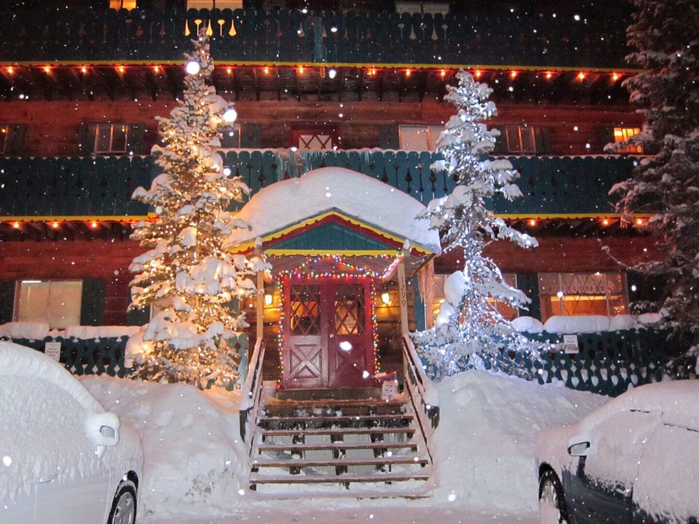 A snow-covered log cabin with lit Christmas trees and festive lights. Snow is falling, and two cars are parked in front.