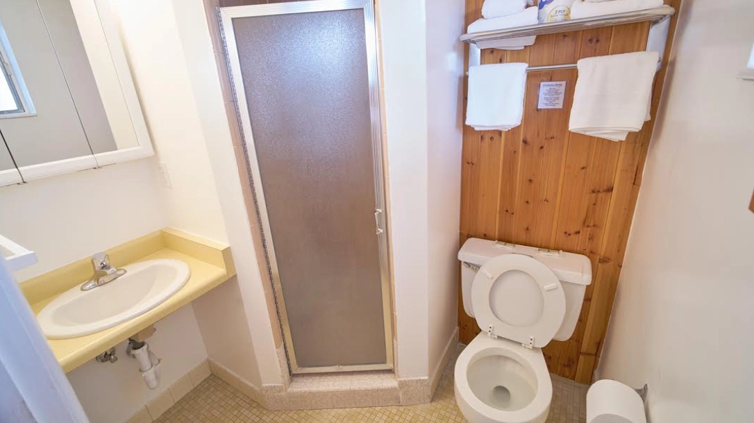 Compact shared dormitory bathroom for skiers featuring a sink, mirror, and shower with sliding door. Wooden paneling adds warmth above the toilet, while towels are conveniently hung for easy access after a long day on the slopes.