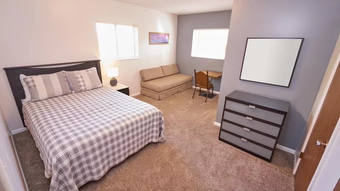 The bedroom, with its dorm-style vibe, features a checkered bed, a nightstand with a lamp, a cozy sofa, a small desk paired with a chair, and a dresser beneath two sunlit windows.