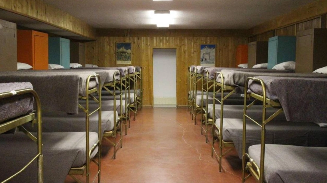 The dorm-style room features rows of metal bunk beds draped in gray blankets, framed by wooden walls and a concrete floor. It's well-lit with ceiling lights.