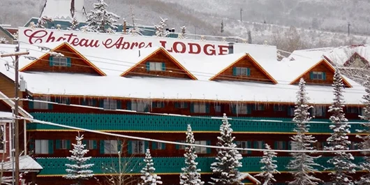 Chateau Apres Lodge in a snowy landscape with several cars parked in front and snow-covered trees around the building.