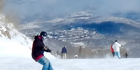 A person gracefully snowboards down the slope, while skiers glide in the background. The stunning snowy landscape and majestic mountains unfold beneath a cloudy sky, perfect for those seeking thrilling adventures followed by cozy evenings in nearby private room rentals.