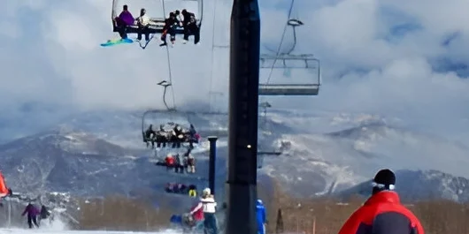 Skiers and snowboarders gracefully carve down the snowy slope as several people enjoy the chairlift in the background. Majestic mountains and clouds provide a stunning backdrop, perfect for those seeking nearby private room rentals to complete their alpine adventure.